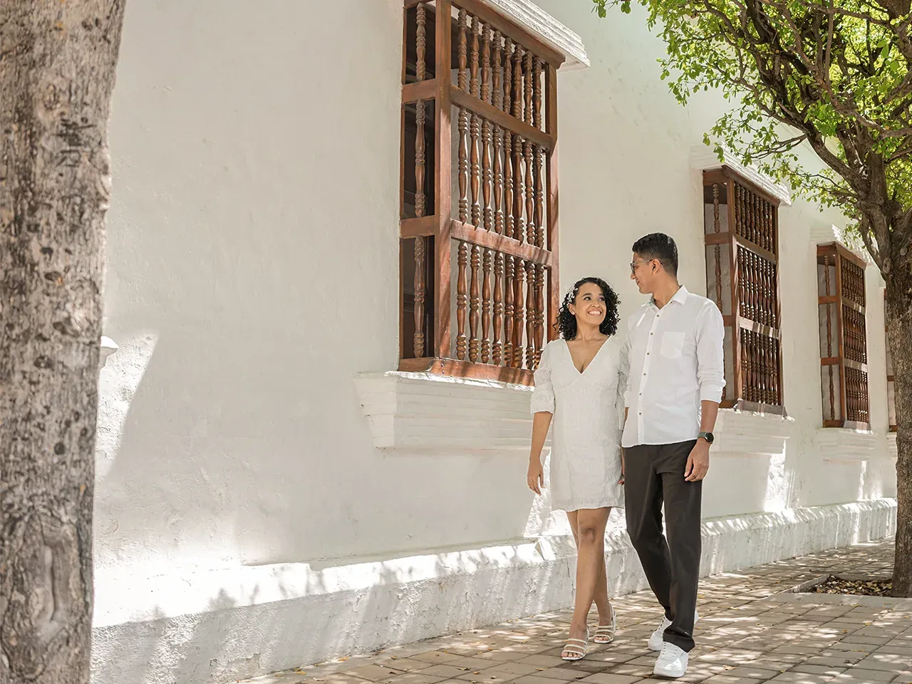 Brian y Lina caminando junto a una fachada blanca con ventanas de madera
